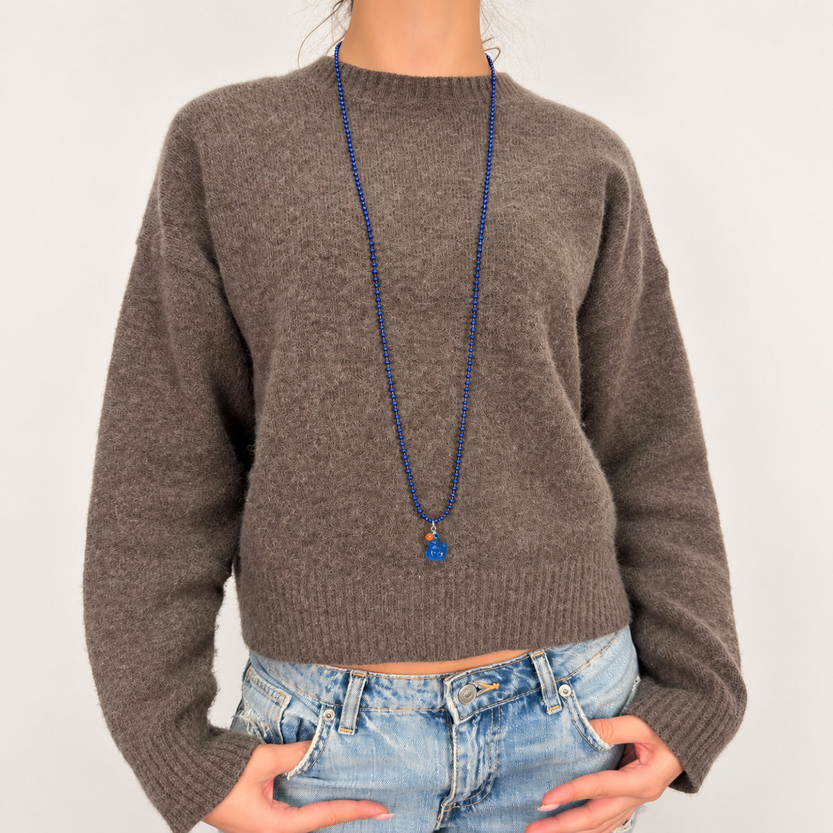 Lange dunkelblaue Kugelkette mit blauem fluoreszierendem Entenanhänger von Escapulario, getragen zu Strickpullover und Jeans.