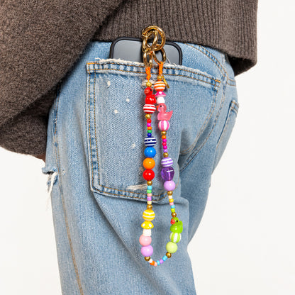 Bunte kurze Handykette von Escapulario mit farbenfrohen Perlen, Entchen und Pilz-Anhängern an einer hellblauen Jeans mit Handy in der Gesäßtasche.