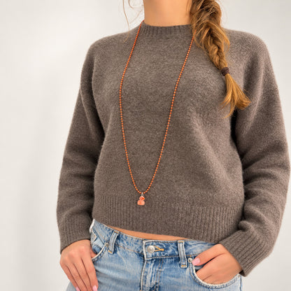 Orangefarbene Metall-Kugelkette mit fluoreszierendem Entenanhänger von Escapulario, kombiniert mit graubraunem Strickpullover und Jeans.