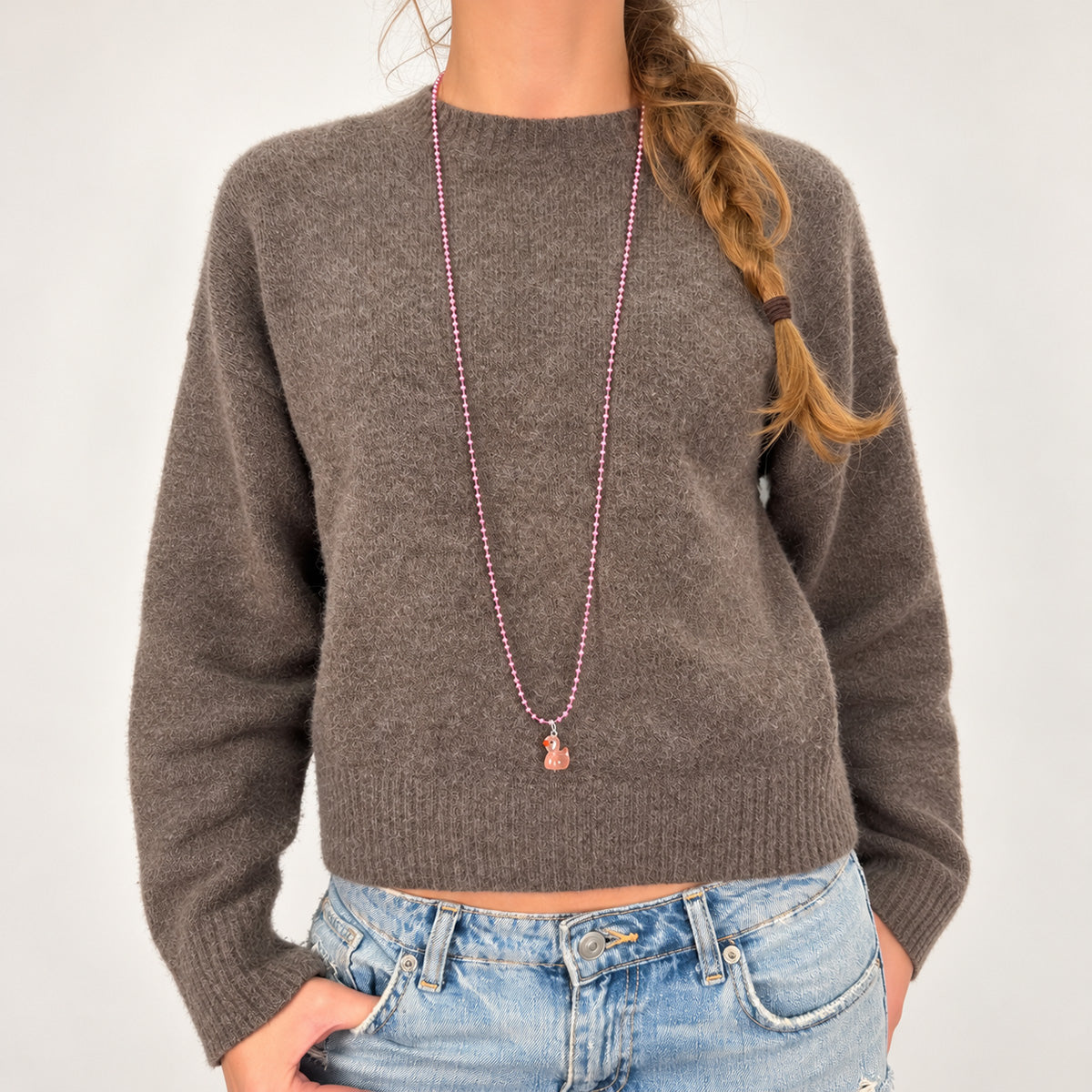 Rosafarbene Metall-Kugelkette von Escapulario mit zartrosa Entenanhänger, kombiniert mit graubraunem Pullover und hellblauer Jeans.