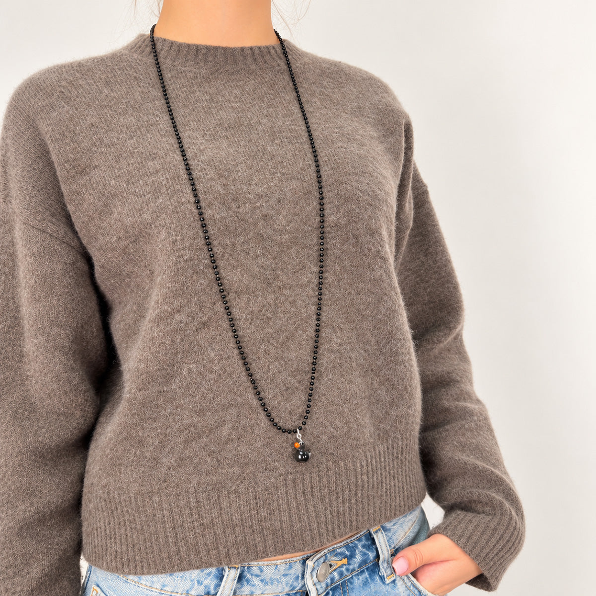 Schwarze Metall-Kugelkette von Escapulario mit schwarzem Entenanhänger und orangefarbenem Schnabel, getragen über einem graubraunen Strickpullover.