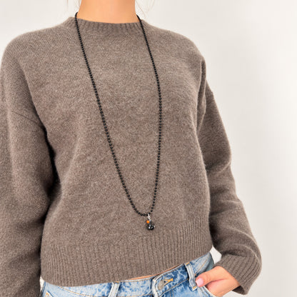Schwarze Metall-Kugelkette von Escapulario mit schwarzem Entenanhänger und orangefarbenem Schnabel, getragen über einem graubraunen Strickpullover.