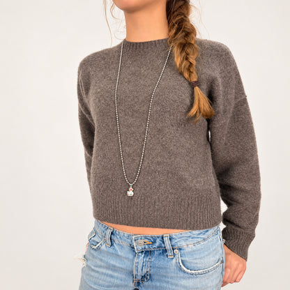 Silberfarbene Metall-Kugelkette von Escapulario mit pinkem Entenanhänger, getragen zu graubraunem Strickpullover und hellblauer Jeans.